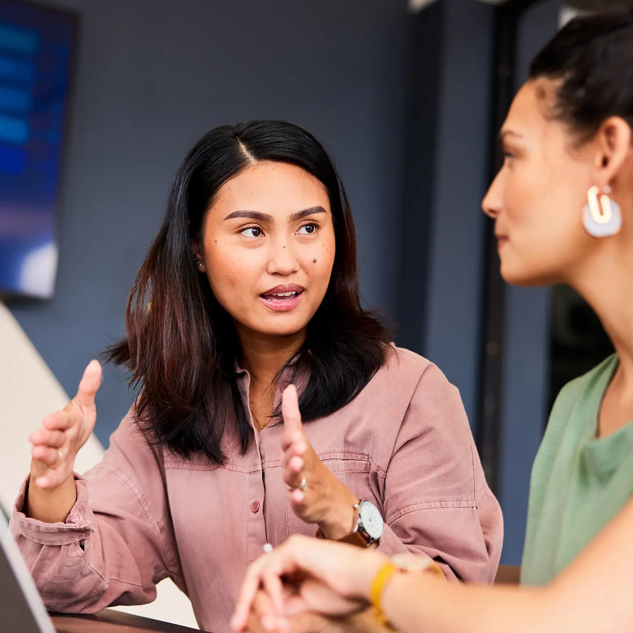 Two women engaged in a discussion at a table, one gesturing with hands. The setting is informal, with a focus on collaboration and communication.