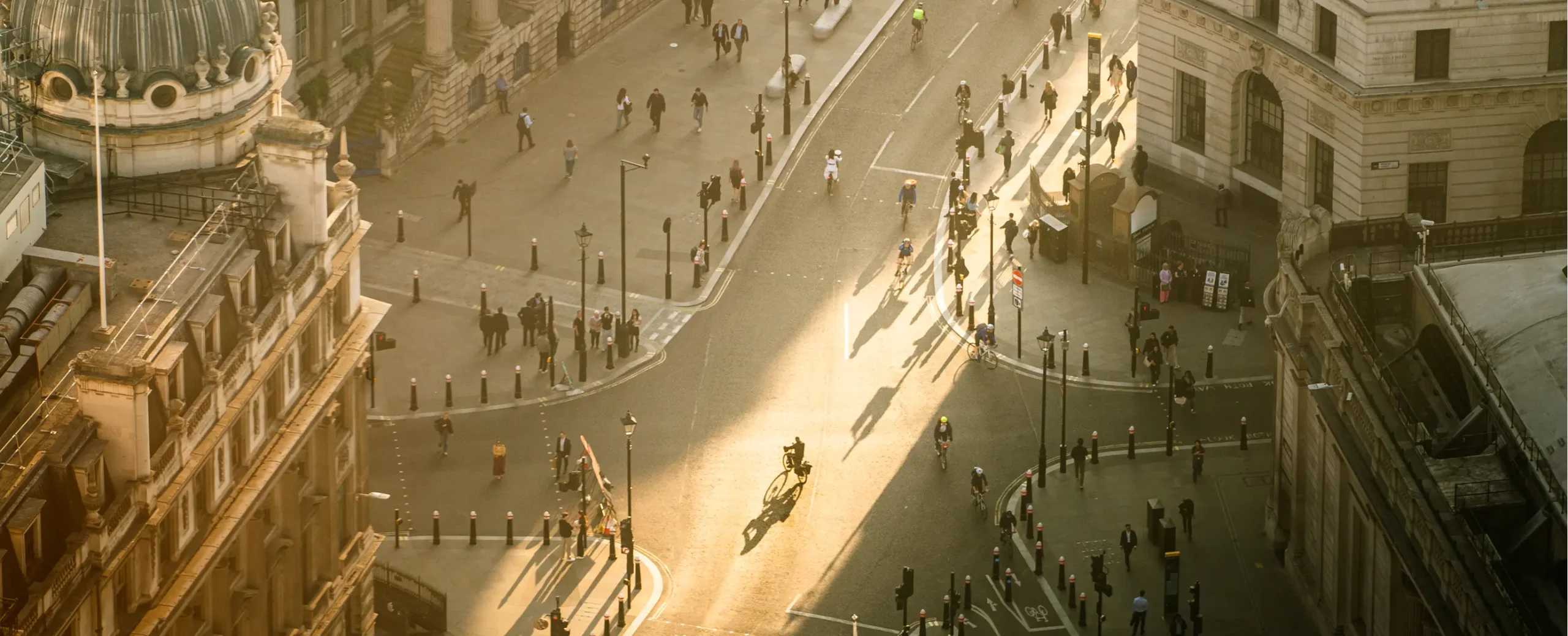 Aerial view of a sunlit city intersection with people walking and a cyclist in the center. Historic buildings surround the scene, creating a warm, bustling atmosphere.