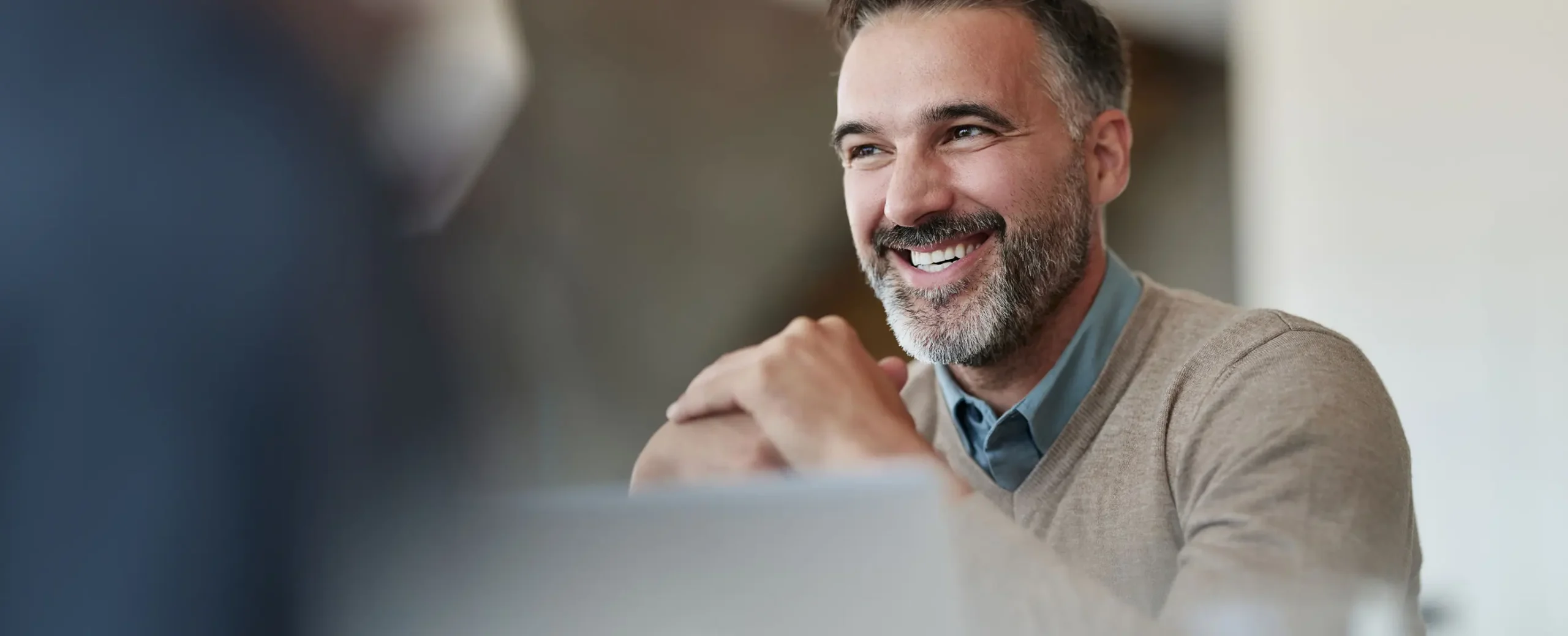 Smiling man with gray hair and beard wearing a beige sweater over a blue shirt, sitting at a table, exuding a friendly and relaxed atmosphere.