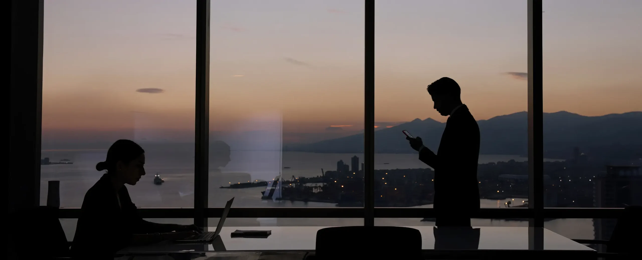 Silhouetted office scene at sunset with a woman using a laptop and a man on a phone, against a window showing a cityscape and mountains. Calm ambiance.