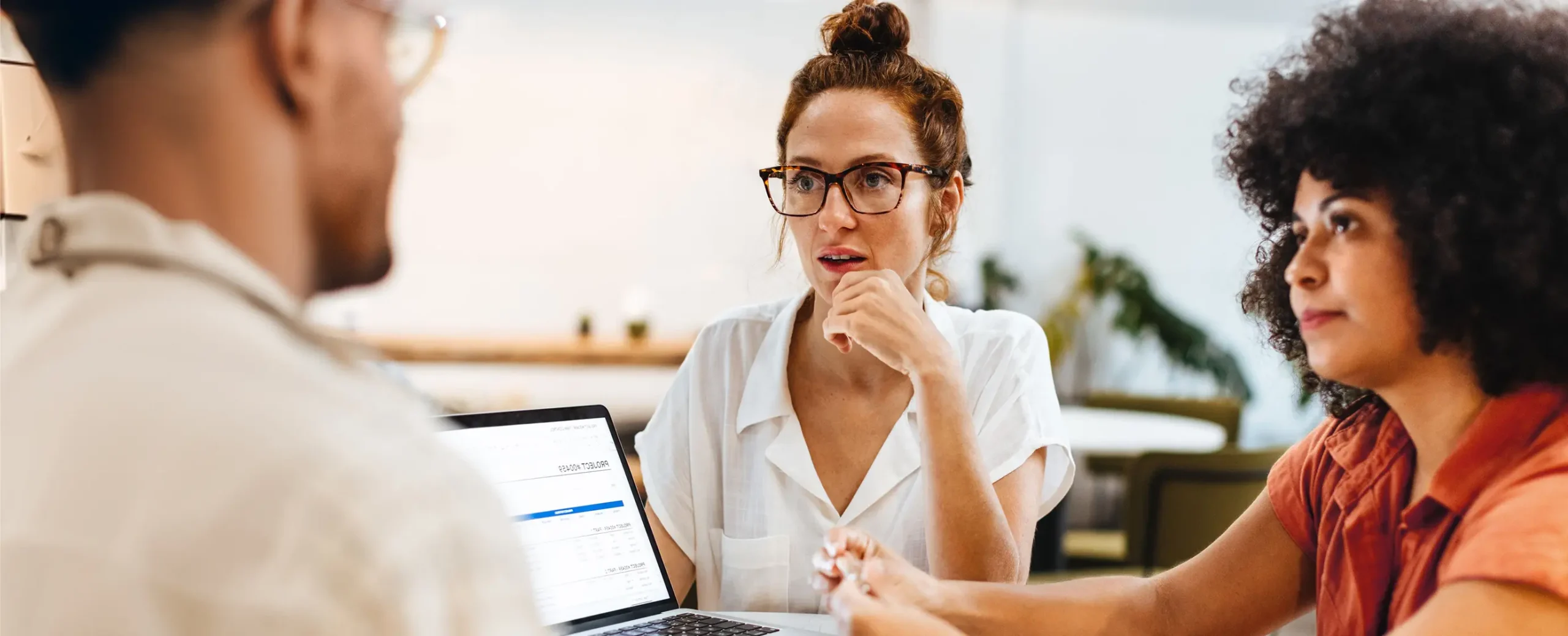 Three people in a casual office setting focus intently on a laptop screen displaying a document. The atmosphere is thoughtful and collaborative.