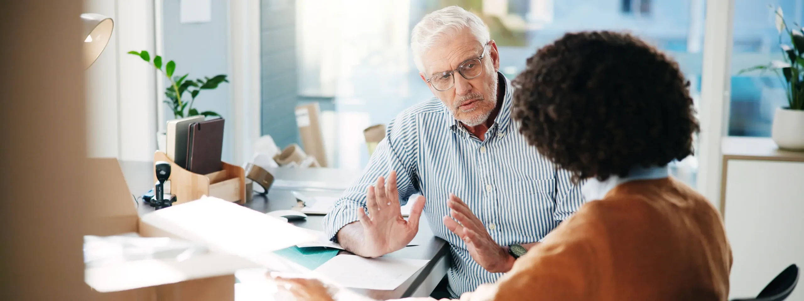 A man with gray hair and glasses gestures while talking to a woman with curly hair in an office. The setting is bright, conveying a focused atmosphere.