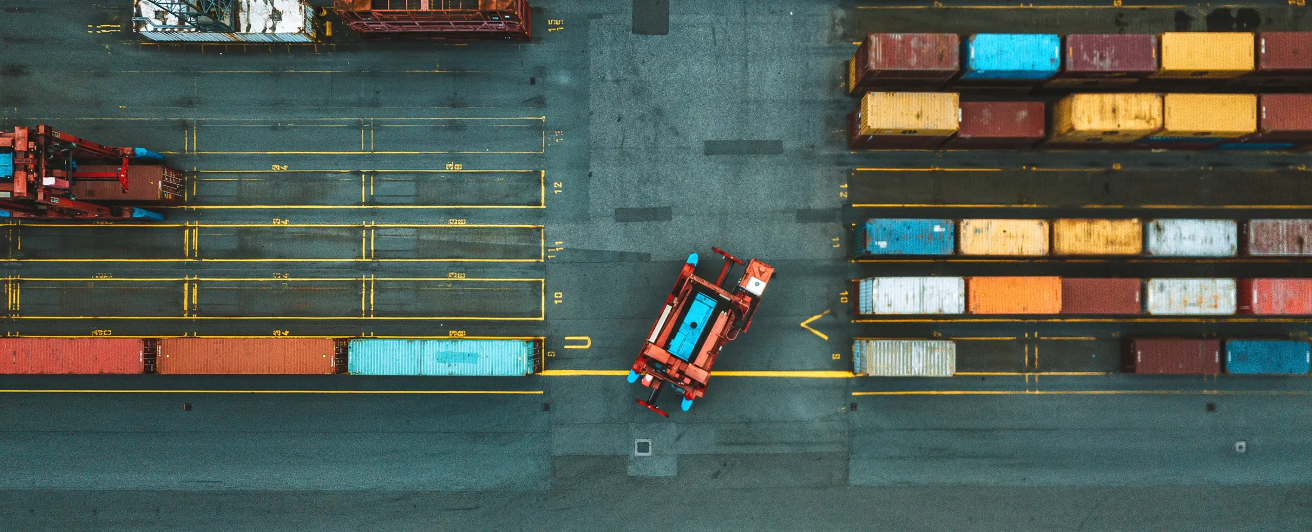 Aerial view of a shipping yard with colorful containers and red trucks on a gray concrete surface, conveying an organized and industrious atmosphere.