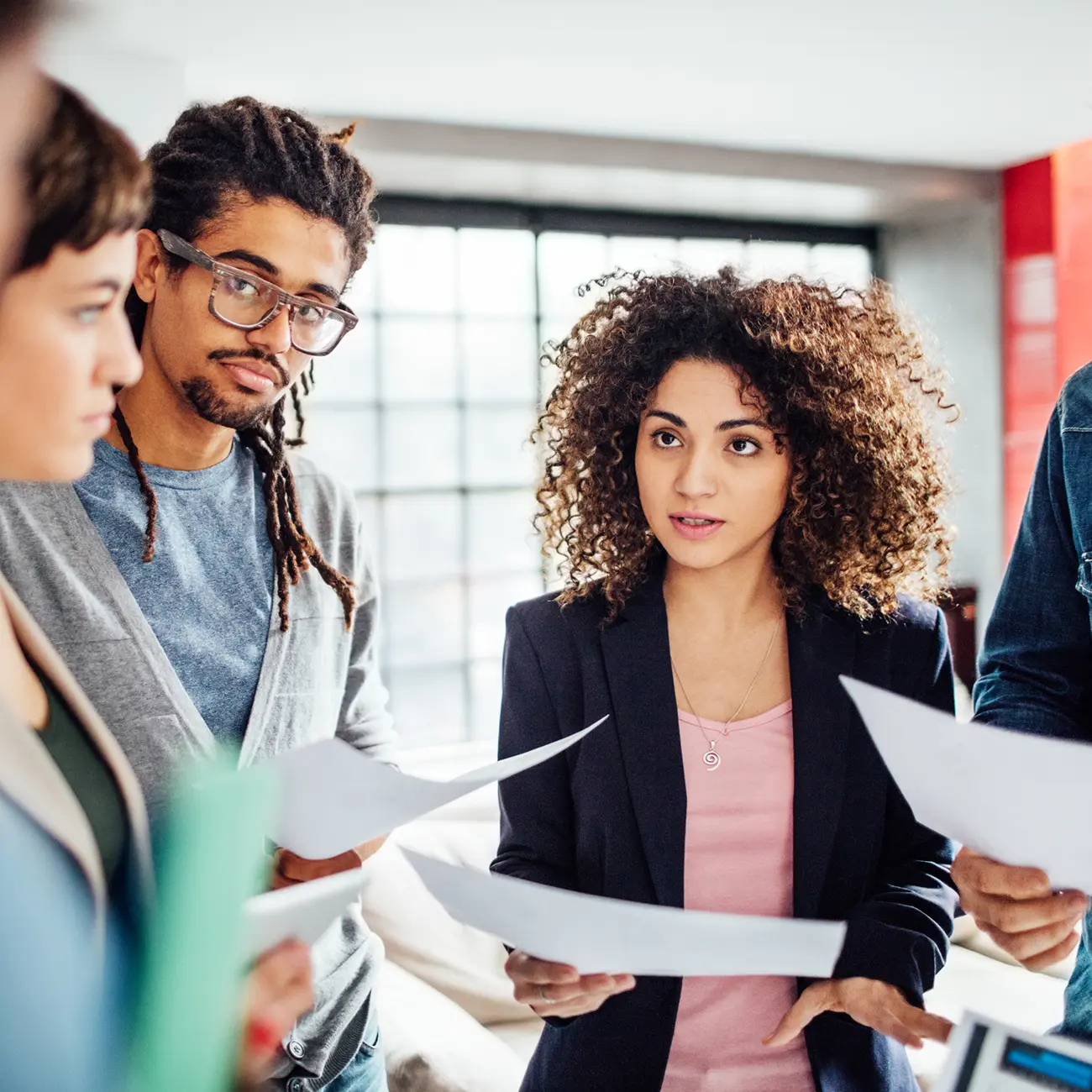 A diverse group of four professionals engaged in a discussion, holding documents. They appear focused and collaborative in a bright office setting.
