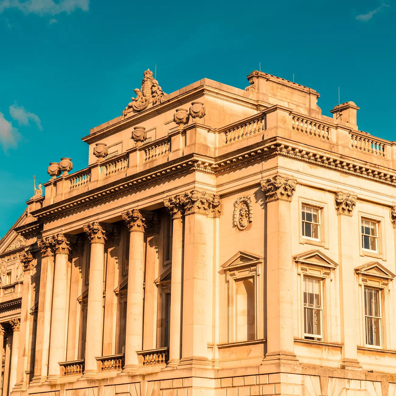 Ornate historic building with classical columns and decorative stonework, bathed in warm sunlight against a vibrant blue sky, exuding elegance and grandeur.