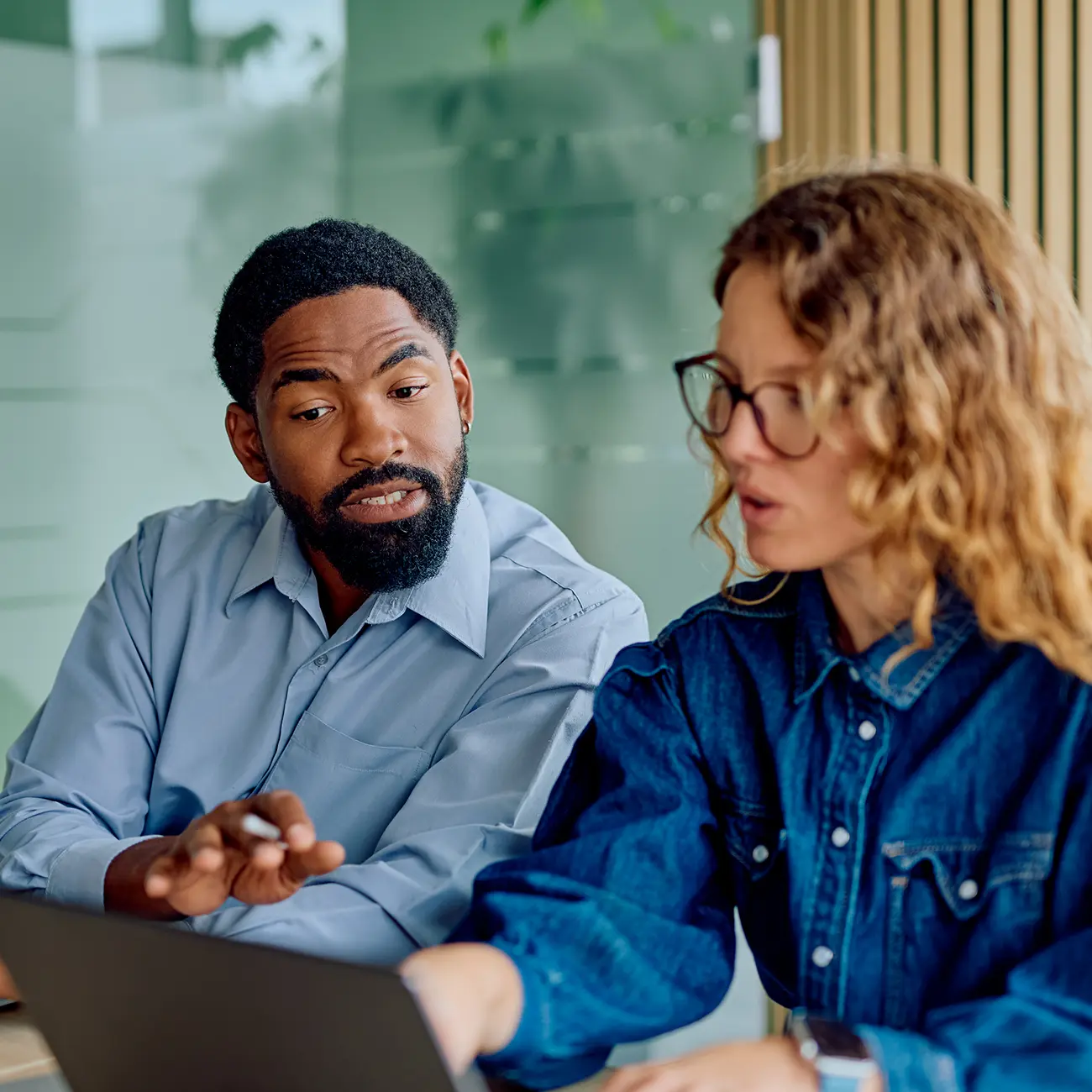 Two coworkers collaborating and discussing a project while working on a laptop in a modern office.