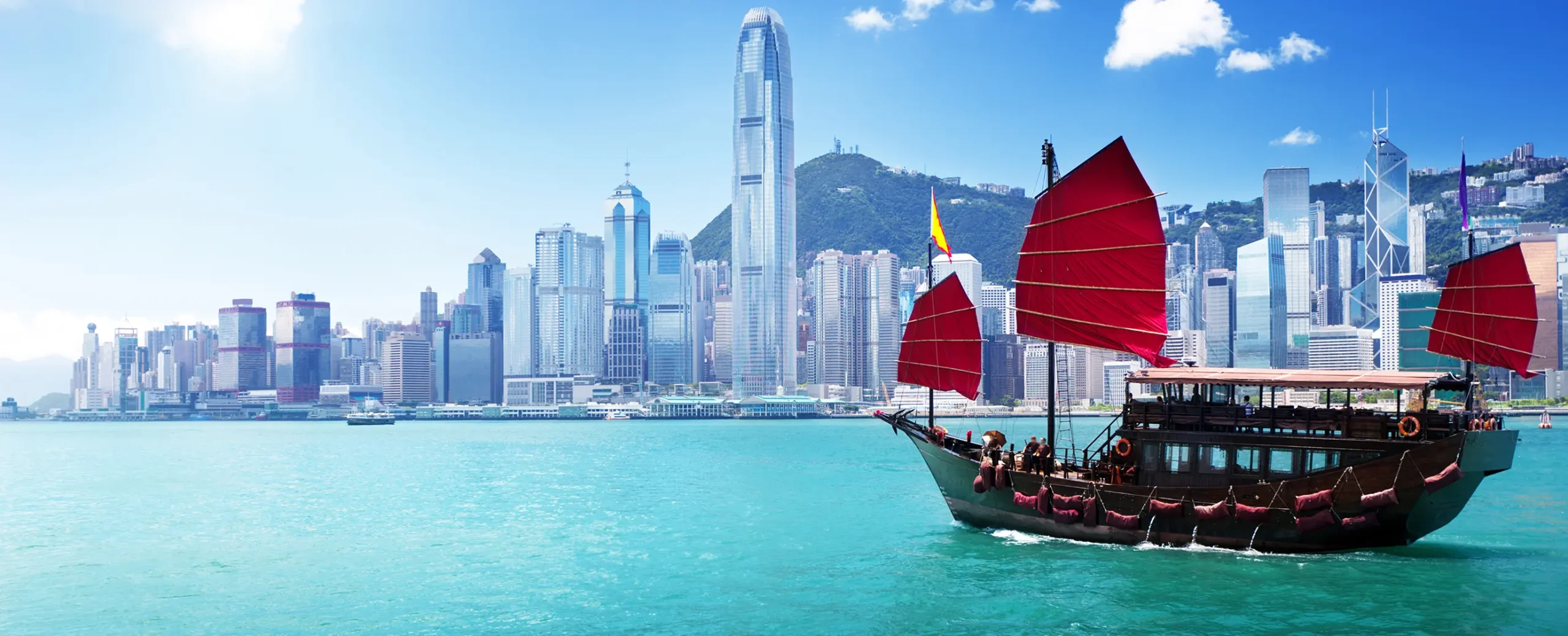 A traditional junk boat with red sails floats on turquoise water against a backdrop of Hong Kong's modern skyline under a clear blue sky.