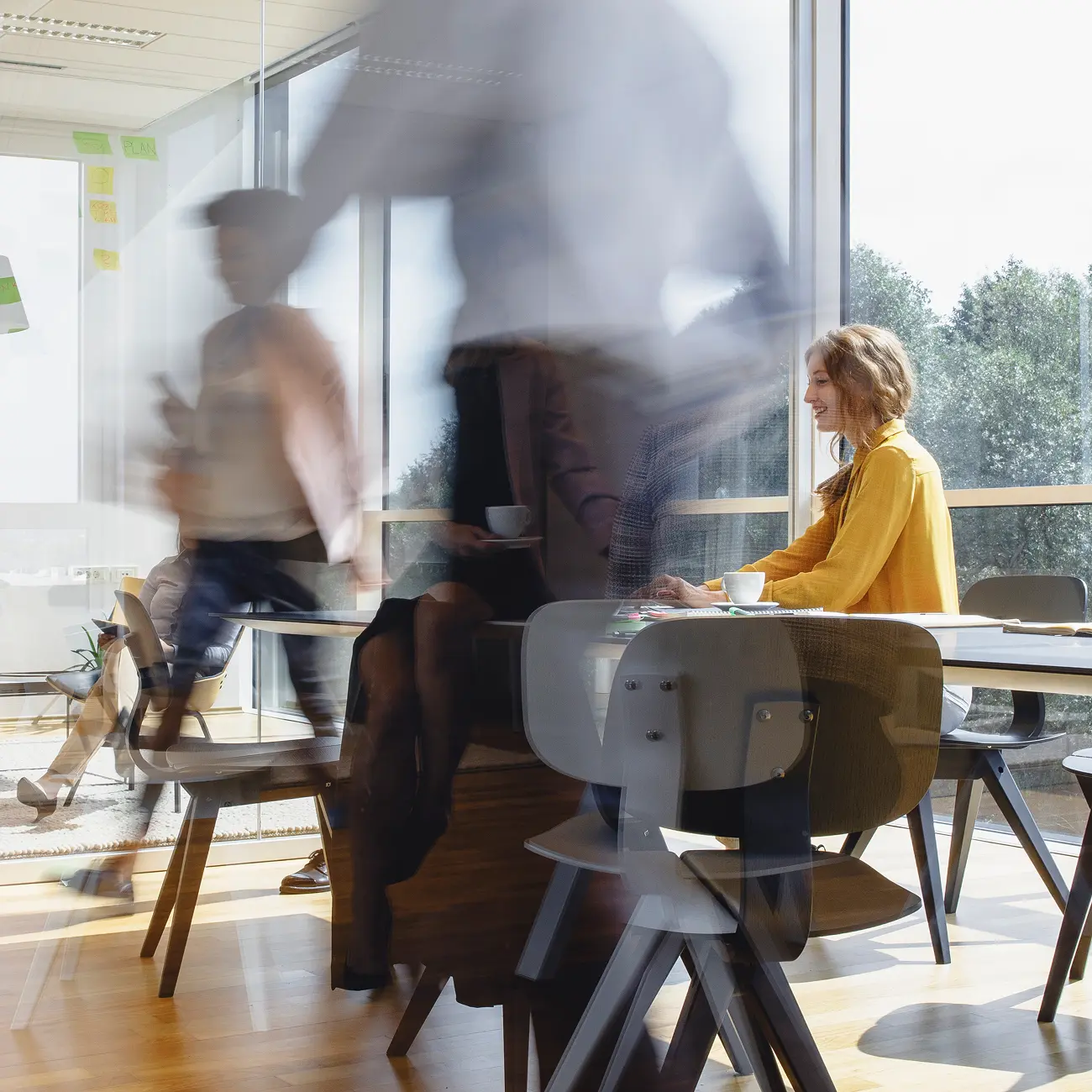 A woman in a yellow shirt sits at a table in a sunlit office, smiling. Blurred figures move around, creating a sense of busy, dynamic activity.