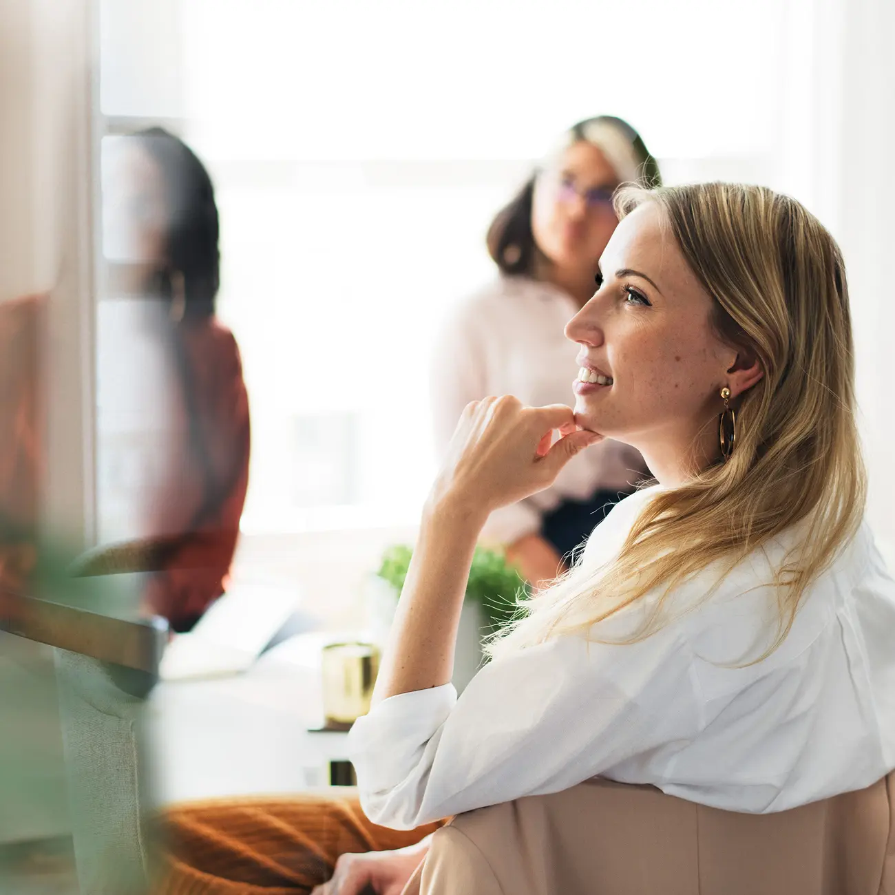 A woman with long blonde hair is smiling thoughtfully, resting her chin on her hand. Two blurred figures in the background create a serene, reflective atmosphere.