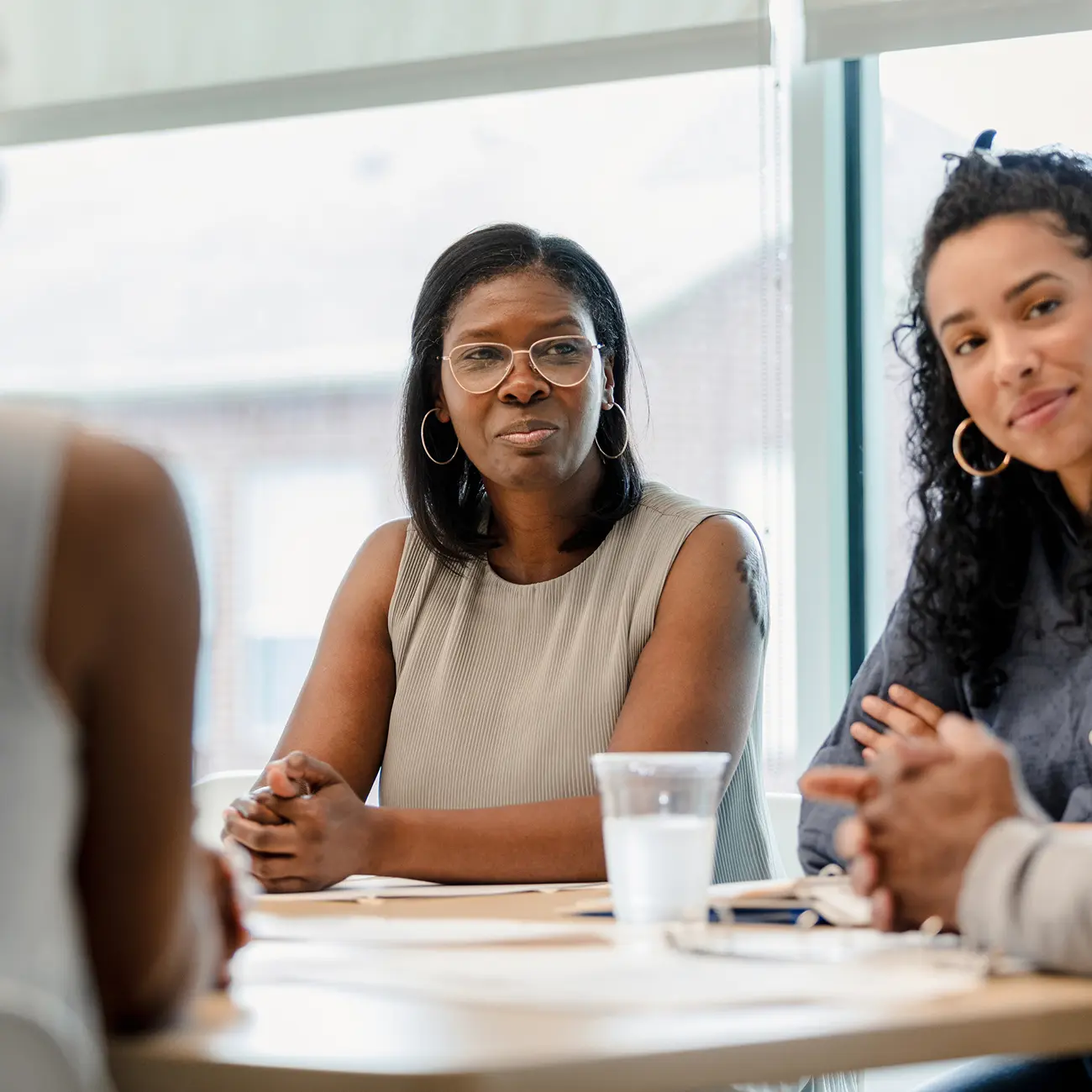 Three people sit around a table in a well-lit office. A woman wearing glasses and a sleeveless top looks attentively. Two others are partially visible.