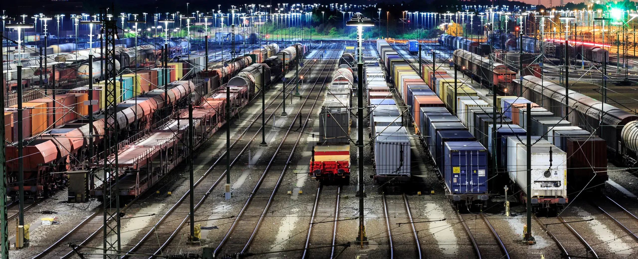 Train yard at night, filled with rows of colorful freight cars under bright lights. Tracks create symmetrical lines, evoking a sense of order and activity.