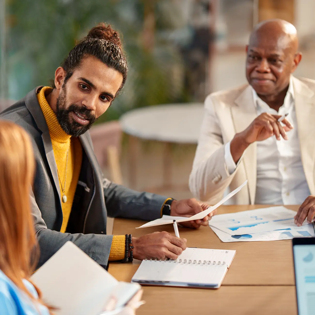 A man with a bun and yellow sweater engages in discussion with two colleagues at a table, holding papers and a notebook, suggesting teamwork in an office setting.