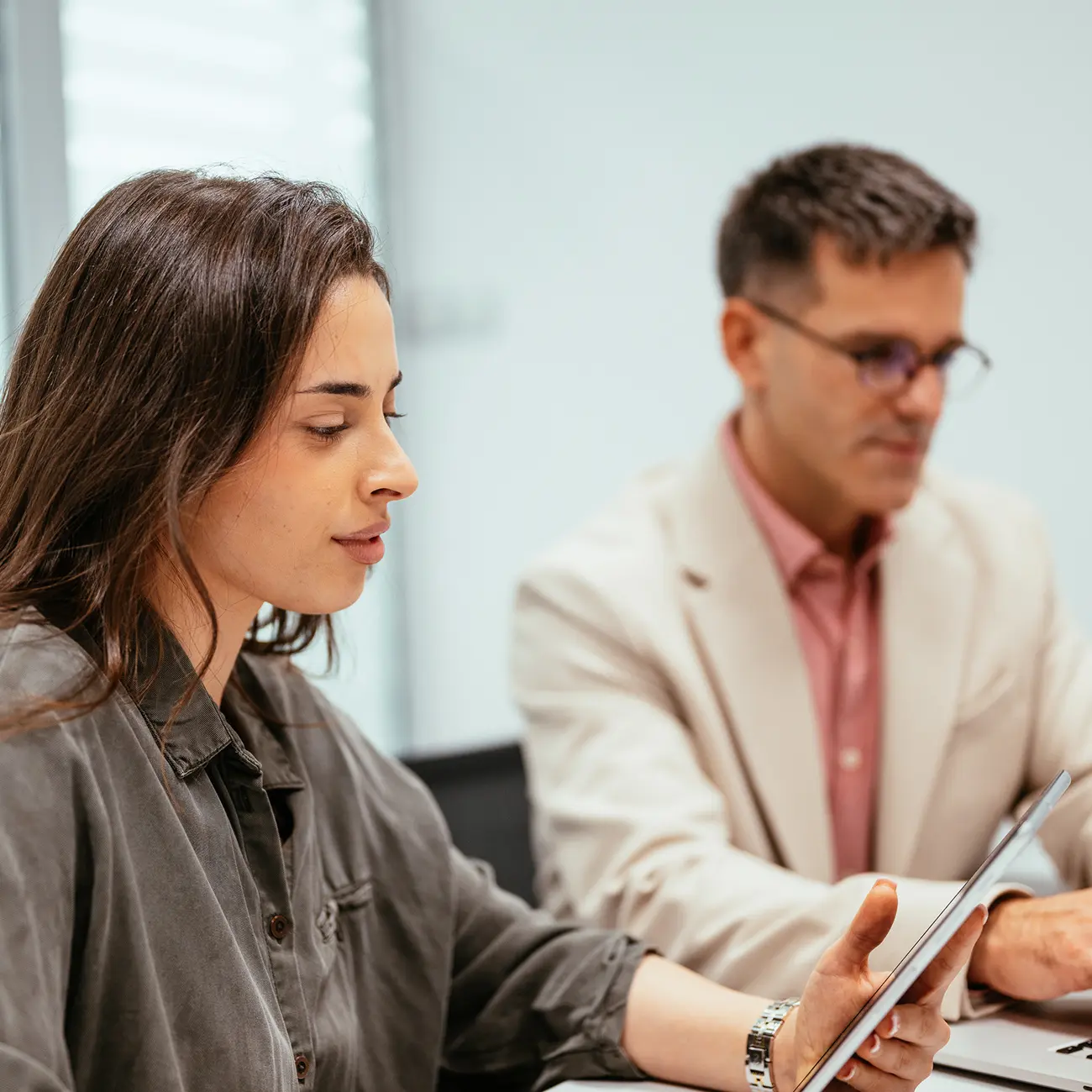 A woman and a man sit at a table focused on a document. The woman wears a gray blouse; the man, blurred, wears glasses and a beige blazer, creating a professional tone.