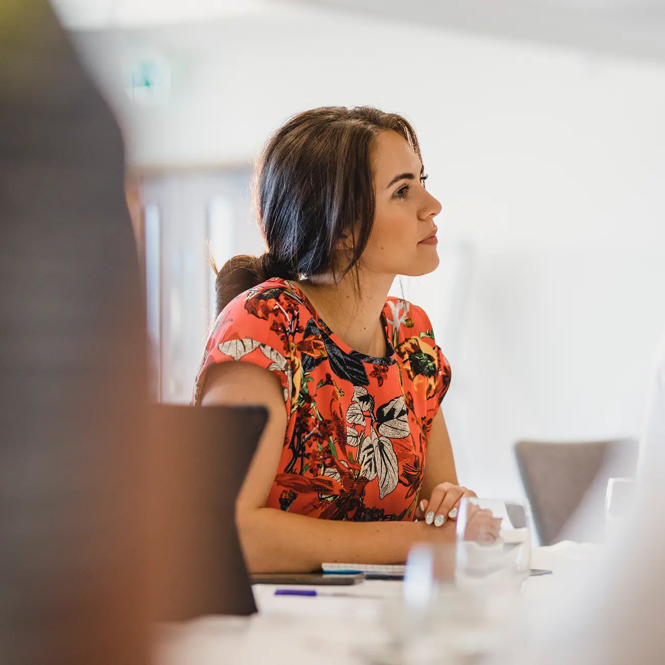Woman in a floral red dress attentively listens during a meeting, leaning forward with hands clasped. Bright, professional setting conveys focus.